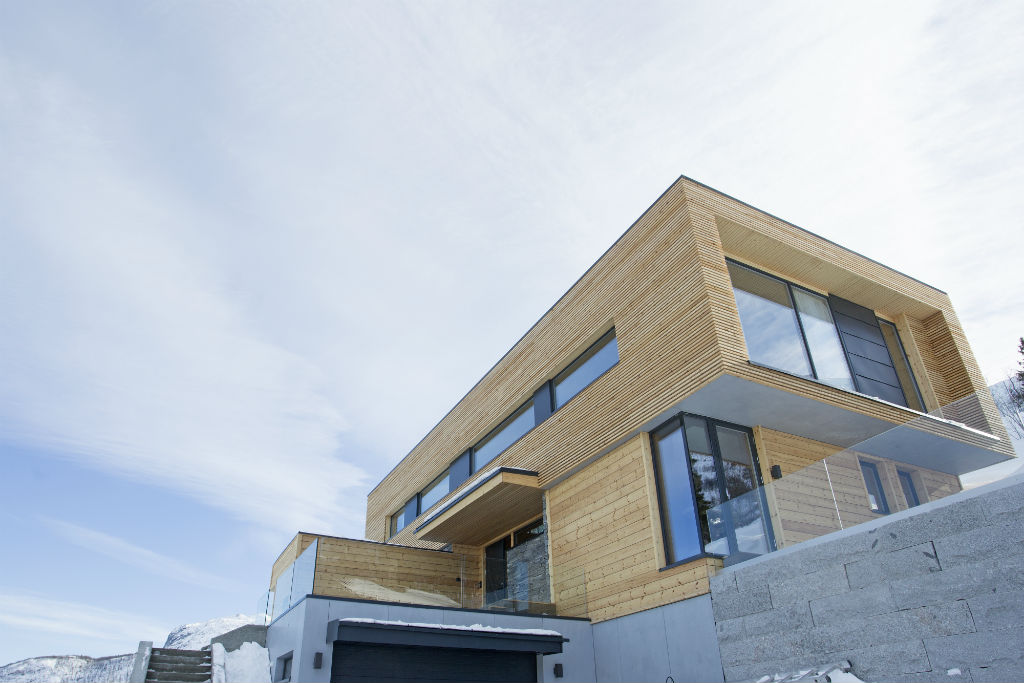 Moderne trekledd hus med store vinduer, betonggrunn, snø og blå himmel.