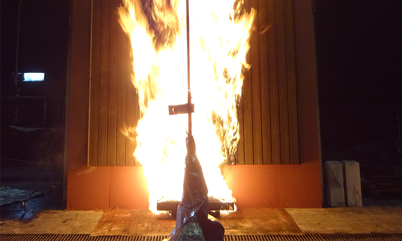 Mørk skinke henger på krok foran intens oransje-gul flamme mot trevegg