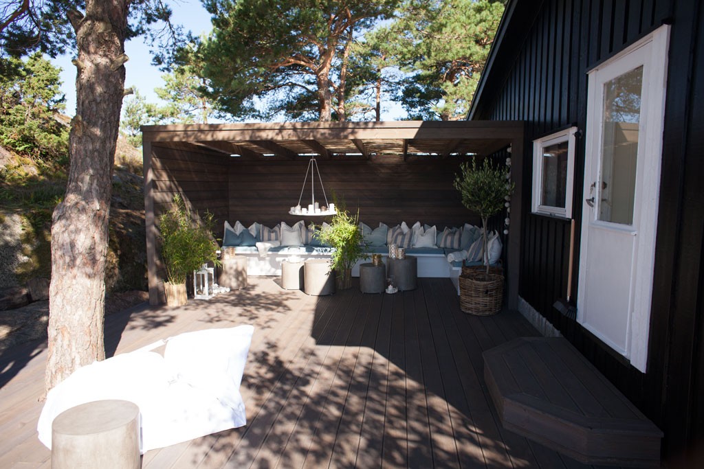 Sort hytte med pergola over sittegruppe på mørkt tregulv, hvite puter og grønne planter.