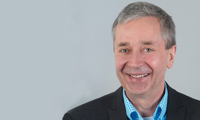 Mann med grått hår iført mørk dressjakke og blå skjorte, stående foran lys grå bakgrunn.