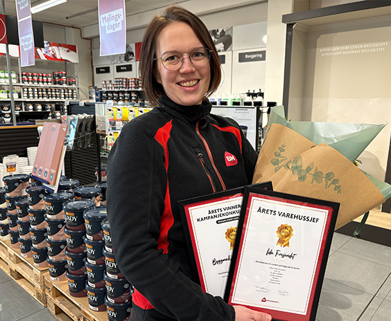 Ansatt i svart-rød uniform holder to diplomrammer og bukett foran hyller med malingsbokser.