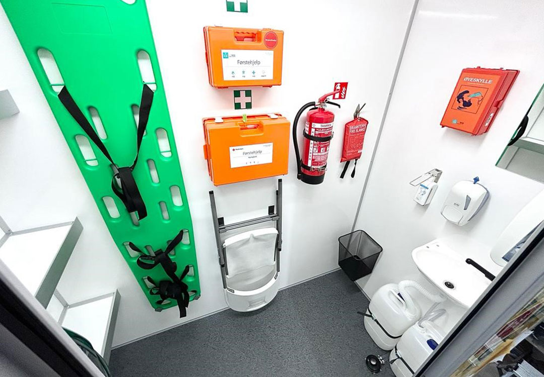 Lite hvitt rom med grønt ryggbrett, oransje førstehjelpskofferter, rød brannslukker, toalett