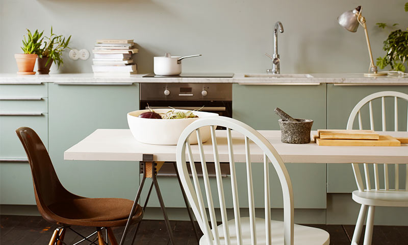 Lys kjøkken med mintgrønne skap, hvitt bord, mismatchede stoler, fruktskål og morter.