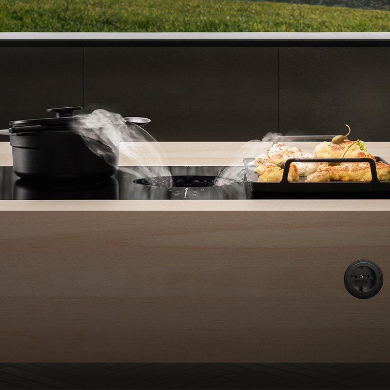 Lys tres benk med svart induksjonstopp, integrert avtrekksvifte, damp fra gryte og bakt blomkål