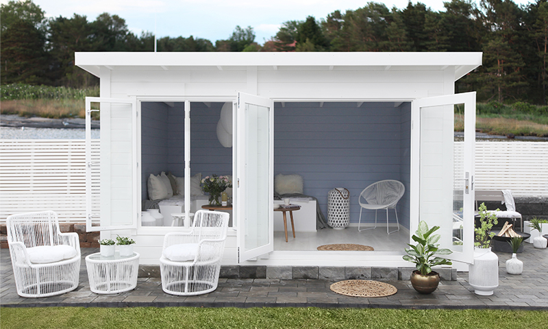 Hvitt hagehus ved sjøen med åpne glassdører, blå bakvegg, hvite rottingstoler og planter.