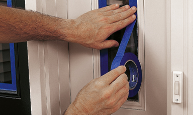 To hender fester blå maskeringstape rundt smalt vindu i beige dør ved dørklokke.