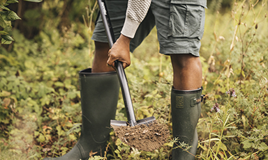 Person i grønne gummistøvler og shorts graver med spade i frodig skogbunn.