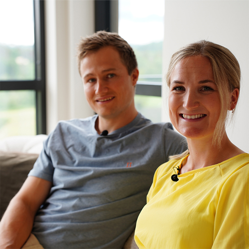 To personer i grå og gul genser sitter på sofa med mikrofonklype foran vindu med grønt landskap