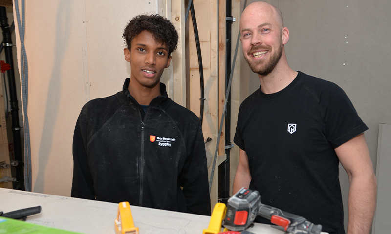 To håndverkere i svart antrekk foran uferdig vegg med trestendere, kabler og verktøy.