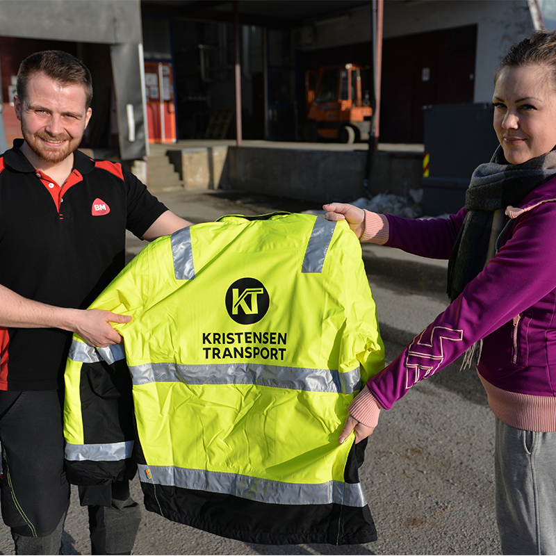 To personer holder gul refleksjakke med svart Kristensen Transport-logo foran lagerbygning.