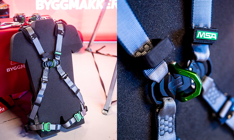 Grått MSA sikkerhetssele med grønne spenner montert på svart torso foran rødt Byggmakker-stand