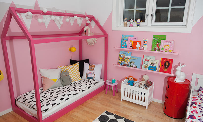 Barnerom med rosa hus-seng, fargerike puter, bokhylle med barnebøker og dukker.