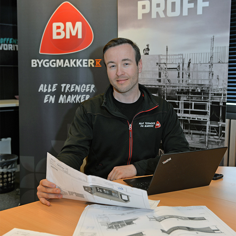Person i svart jakke med røde detaljer, foran Byggmakker-banner, bærbar PC og byggeplaner