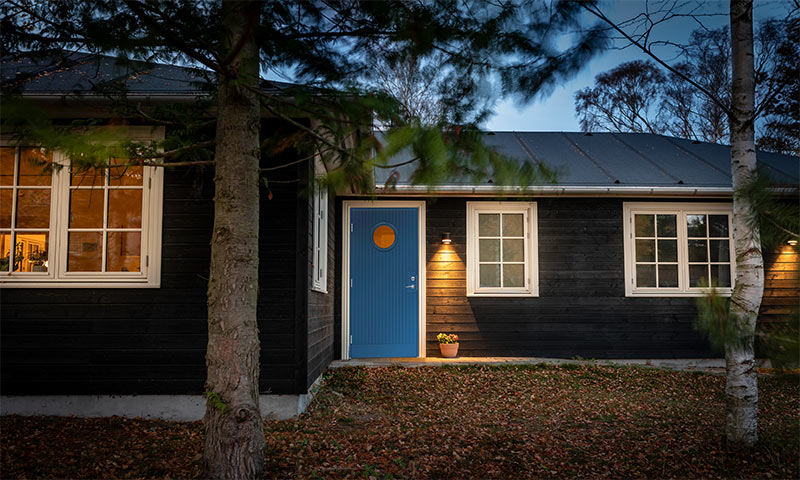 Mørkt trehus i skumring med blå dør og rundt vindu, varme lamper, høstløv på bakken