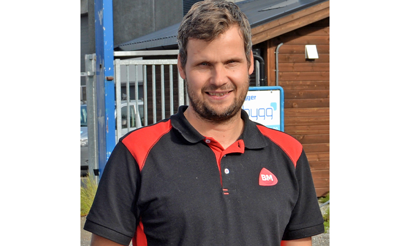 Mann i svart- og rød BM-polo utenfor byggevarebutikk med metallgjerde og trekledning.