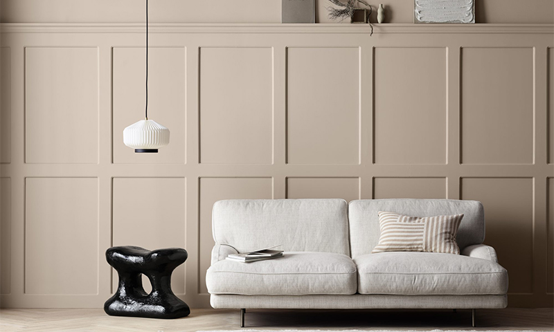 Beige vegg med panel, lys grå sofa med stripet pute, svart skulpturelt bord og hengependel.