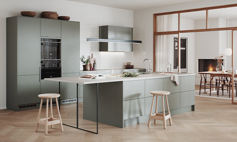 Moderne kjøkken med salviegrønne skap, kjøkkenøy, lyse barkrakker og åpen stue med peis.