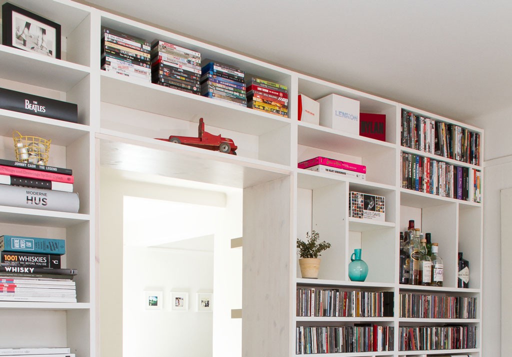 Hvit innebygd bokhylle med fargerike bøker, CD-er, DVD-er, flasker, rød lekebil og plante