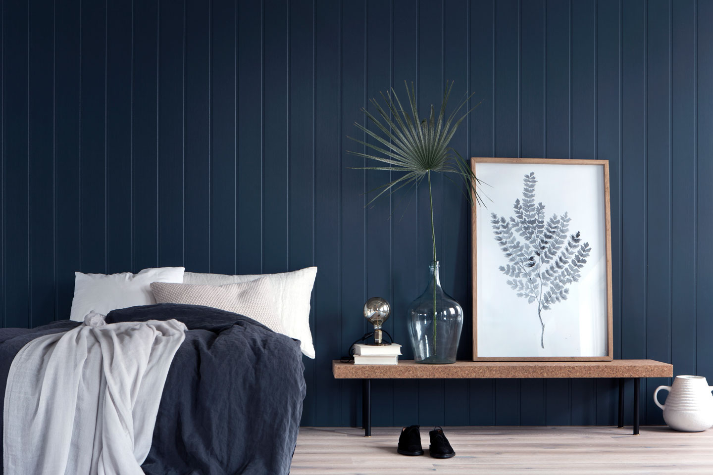 Soverom med mørkeblå panelvegg, seng med blått og lyst teppe, benk med vase, plante og plakat