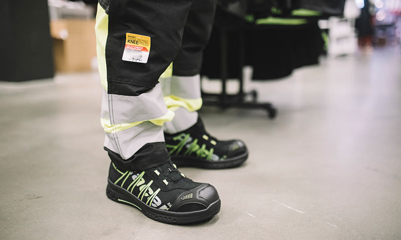 Nærbilde av neongule og svarte arbeidsbukser med knestøtte og svarte vernesko med grønn dekor.