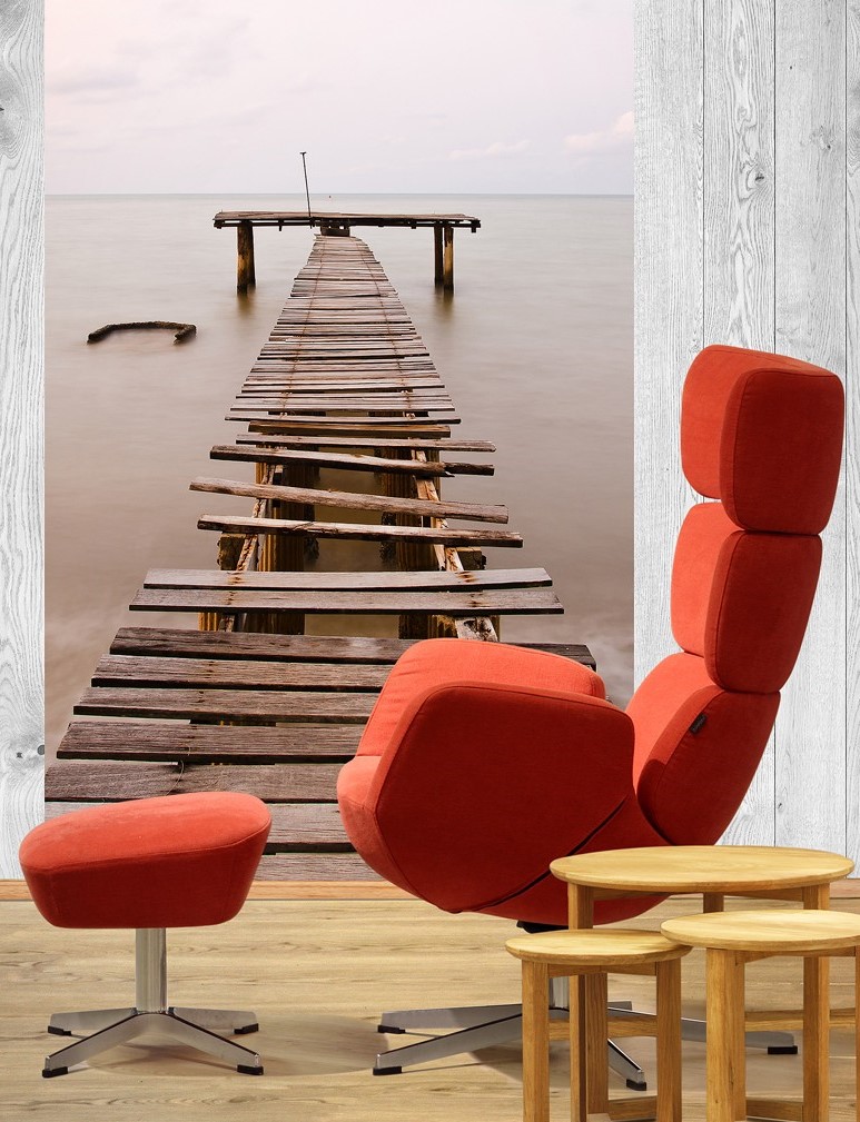 Rød lenestol med fotstøtte og tre små trebord foran bilde av slitt trebrygge mot still sjø