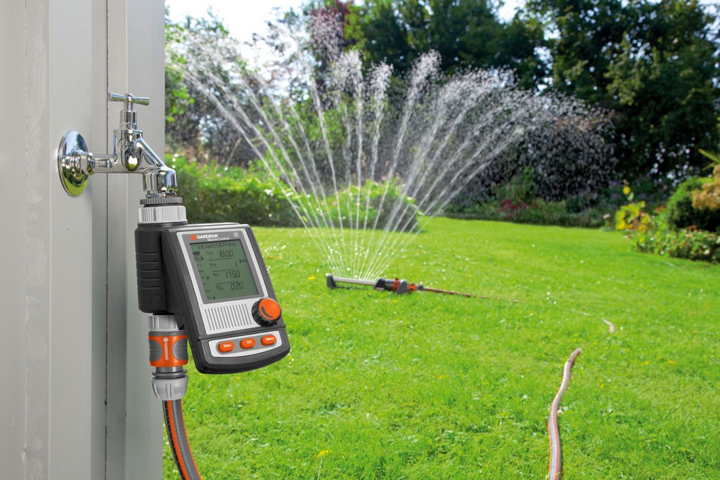 Oransje digital vanningsur på grå utekran med slange, sprinkler sprayer grønt gress