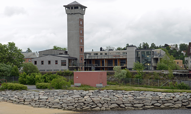 Grå betongbygning med vaktårn og glassfasade ved steinfylt elvebredd under overskyet himmel.