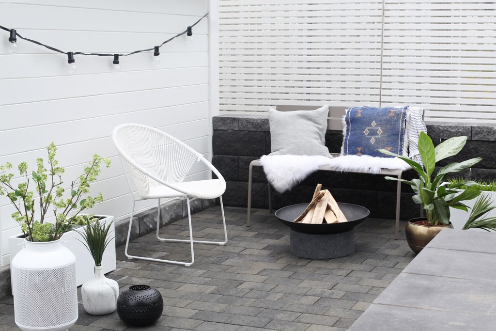 Minimalistisk uteplass med hvit stålstol, benk med grå pute og blått pledd, bålfat og planter.
