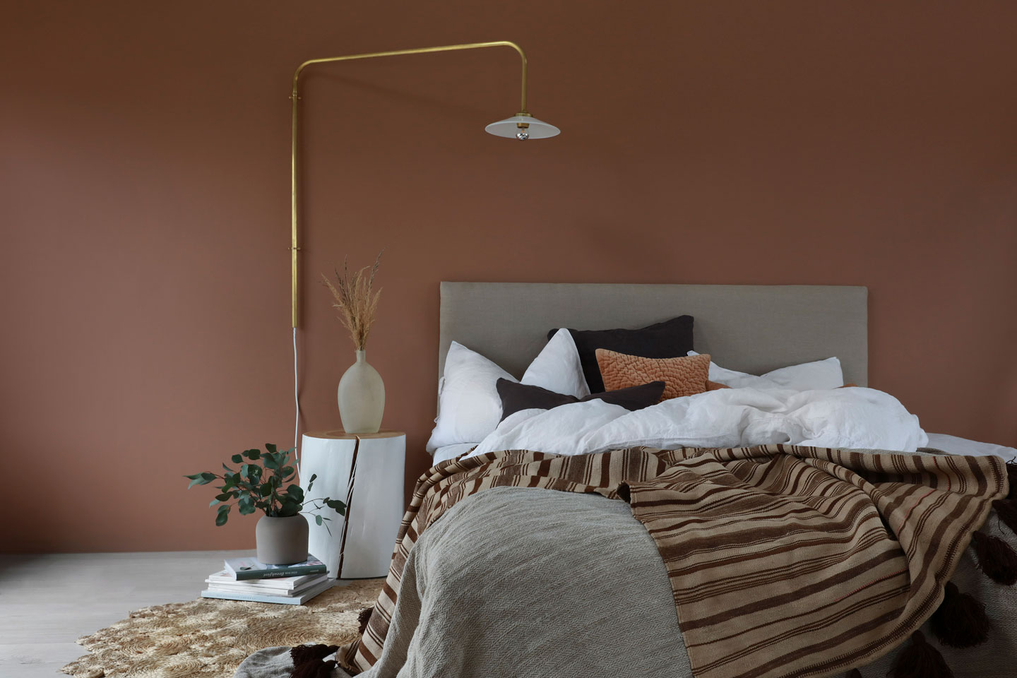 Soverom med terrakottavegg, seng med hvite laken, brune puter, stripete pledd og gyllen lampe