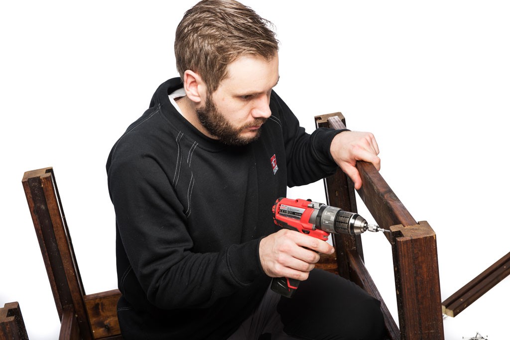 Mann med svart genser bruker rød drill på mørkt trebord mot hvit bakgrunn.