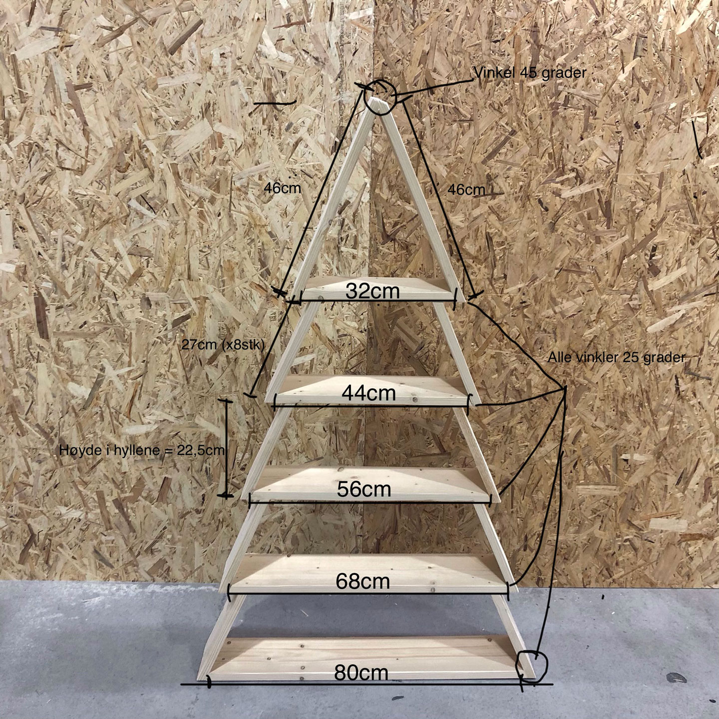 Trepyramidehylle med fem planker mot sponplatevegg på betonggulv, merket med mål.
