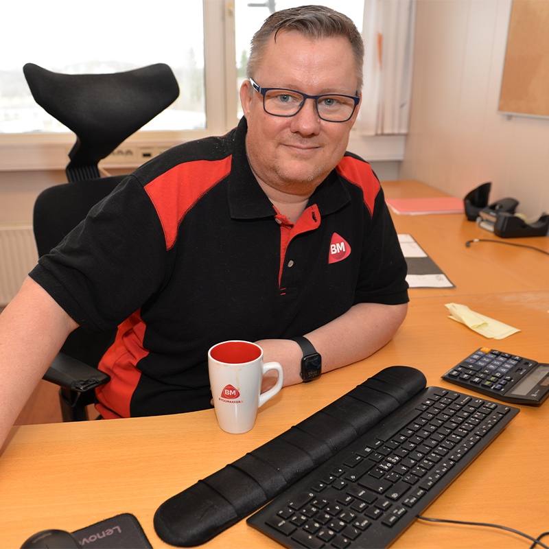 Ansatt i svart-rød BM-polo ved kontorpult med BM-kopp, tastatur og kalkulator.