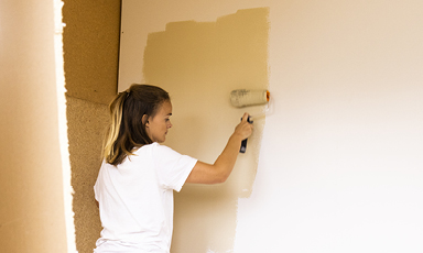 Person i hvit t-skjorte maler innendørs vegg lys beige med malerull.