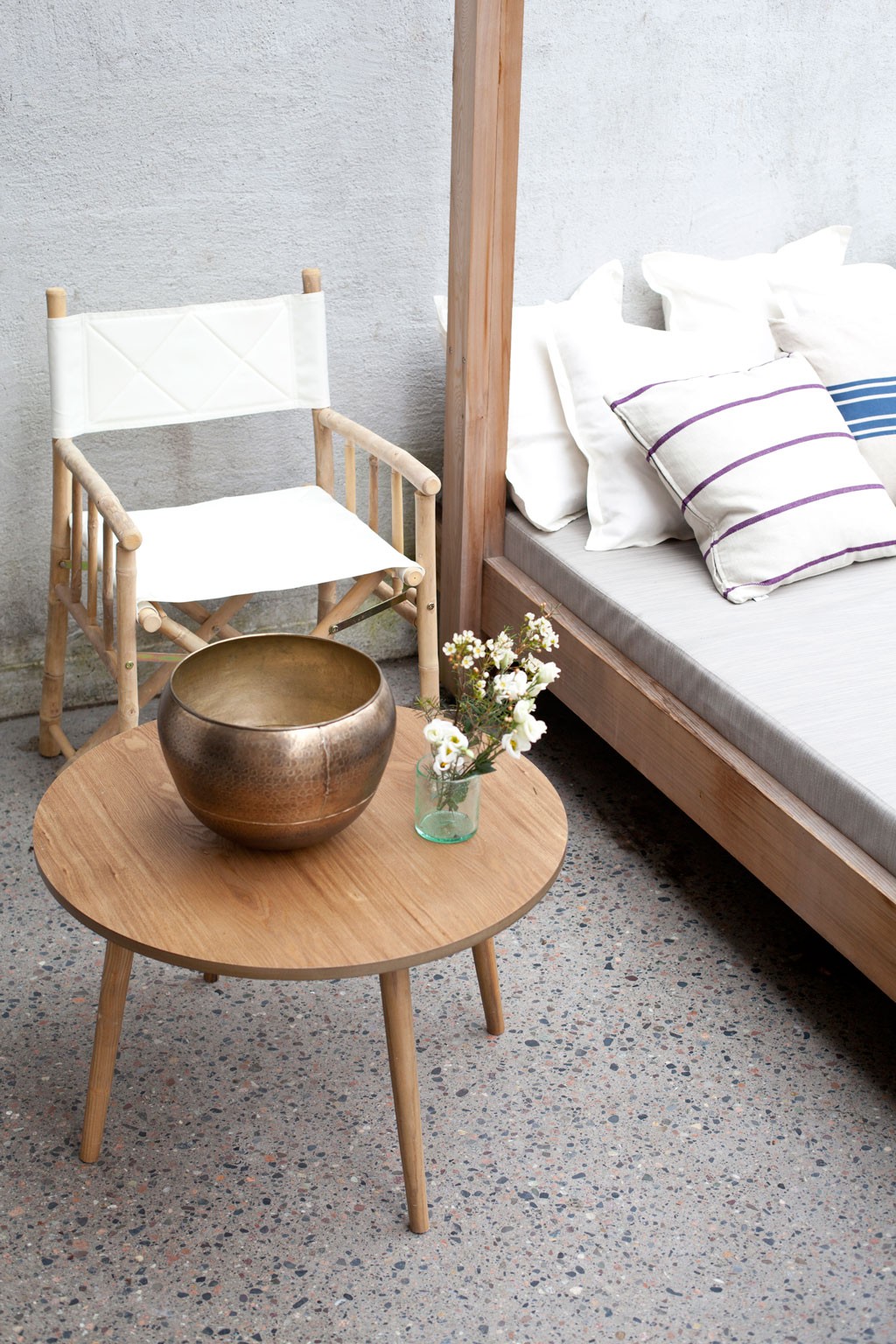 Lyst bord med bronsebolle og små hvite blomster, hvit lerretstol og daybed med stripete puter