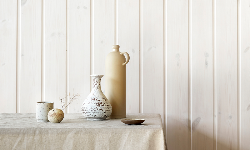 Lyse keramikkvaser og krukke med liten kopp, rund stein og skål på linduk foran lyst trepanel