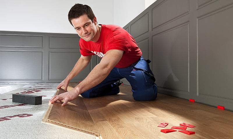 Håndverker i rød t-skjorte og blå bukser legger lys eikeparkett i rom med grå panelvegger.