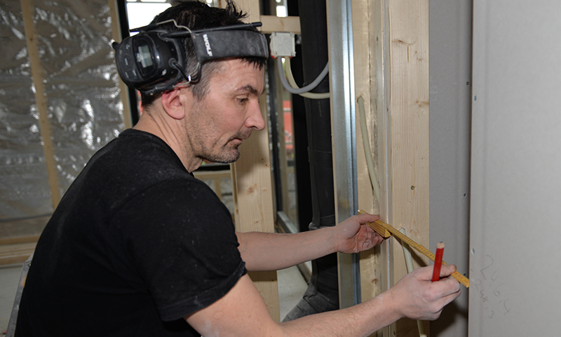 Person i svart t-skjorte måler trestender med gul meterstokk og rød blyant mellom metallstendere.