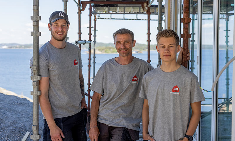 Tre håndverkere i grå BM T-skjorter på stillas ved moderne glassfasade og sjø.