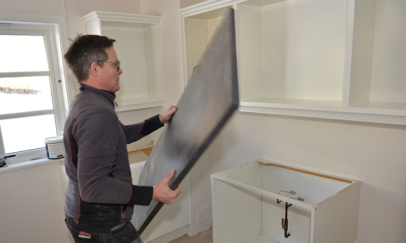 Håndverker løfter svart benkeplate i hvitt kjøkken under montering av skap