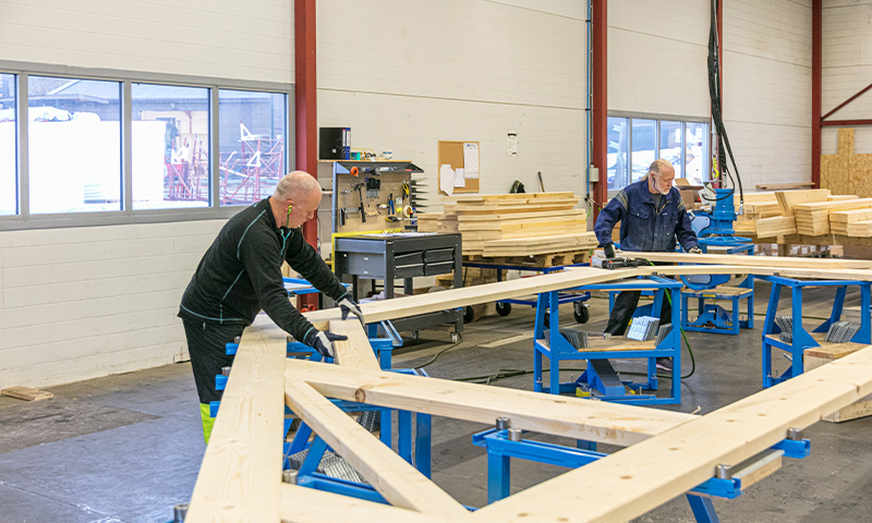 To håndverkere monterer trekonstruksjon på blå stativer i lyst verksted