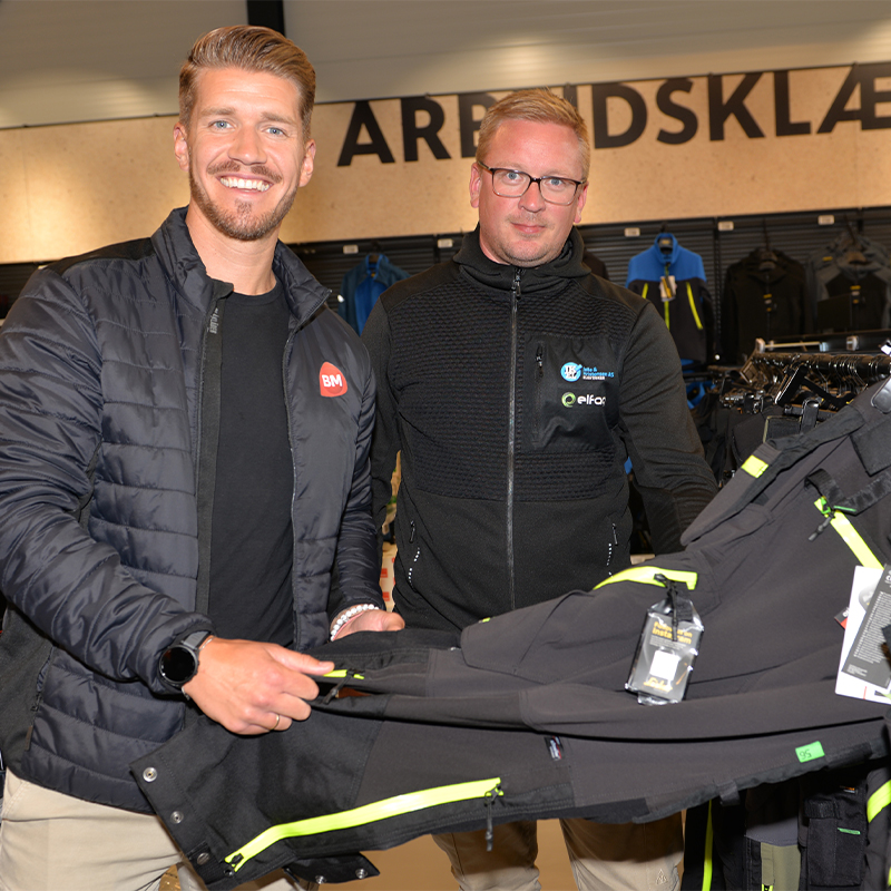 To personer i arbeidsklærbutikk viser svarte bukser med neongrønne glidelåser.