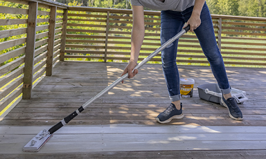 Nærbilde: person i jeans og joggesko maler terrassegulv hvitt med rull, gjerde og grønn hage