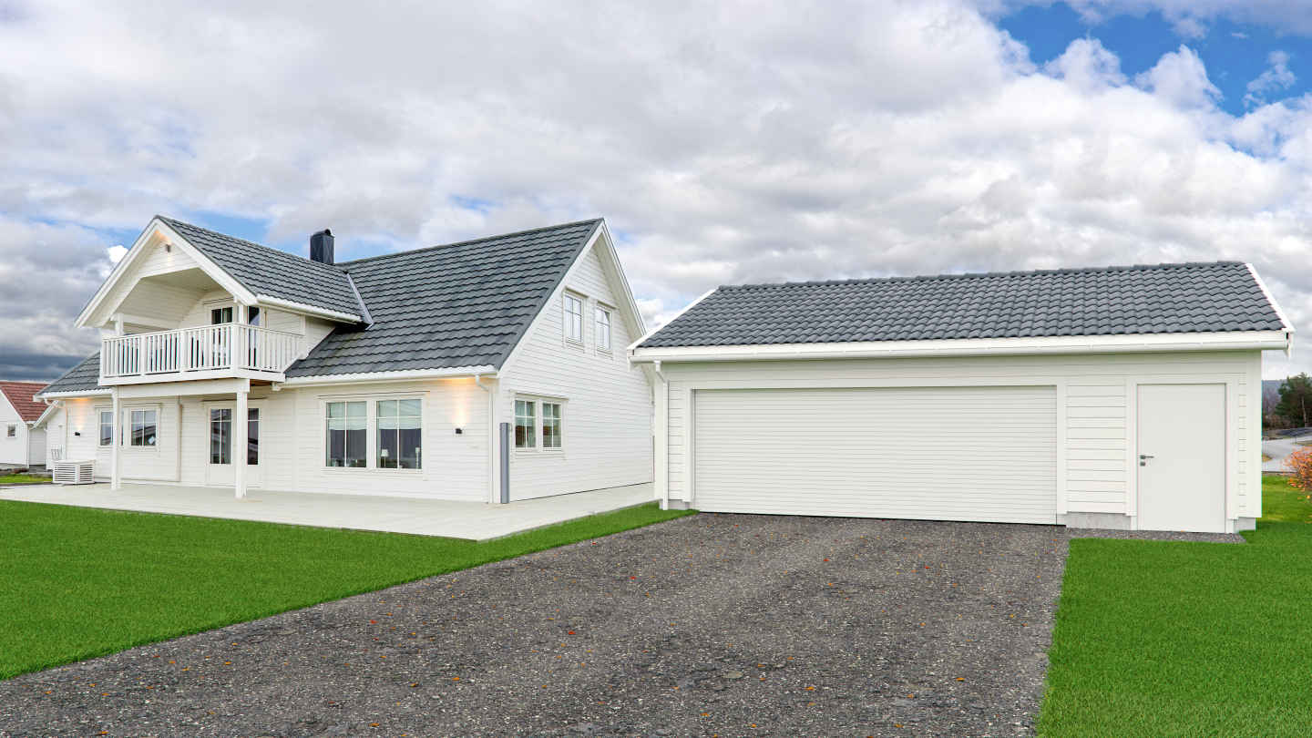 Hvit enebolig med grått tak og balkong, dobbelgarasje, grønn plen og overskyet himmel.