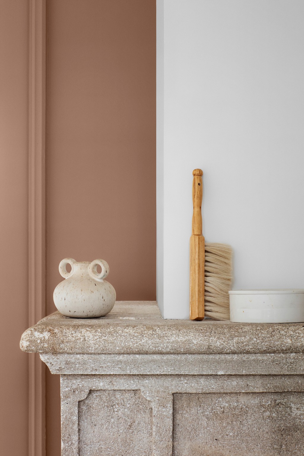 Beigefarget keramikkvase med doble hanker, trebørste og hvit skål mot terrakotta- og hvit vegg.