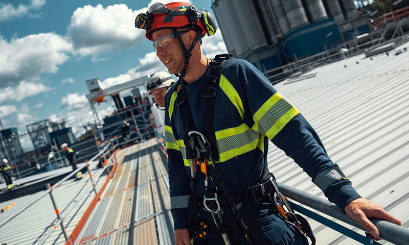 Byggearbeider med rød hjelm, blå drakt og gule refleksstriper på metalltak ved fabrikk