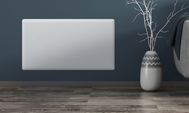 Hvit vegghengt panelovn på mørkeblågrå vegg, tregulv, vase med hvite greiner og grå stol.