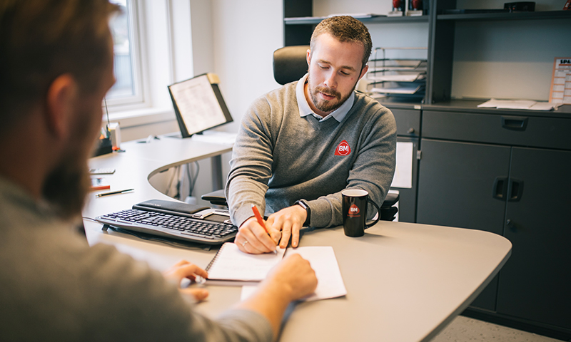 To personer i møte ved lys grå kontorpult med notatblokk, penn, svart BM-kopp og hylle bak.
