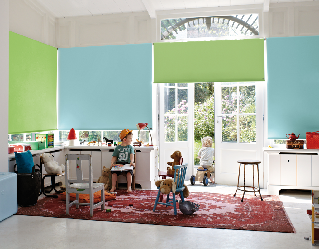 Lyst barnerom med grønne og lyseblå rullegardiner, rødt teppe, leker og hageutsikt.