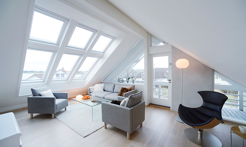 Lyst loftstue med skråtak, store takvinduer, grå sofaer, glassbord, svart lenestol