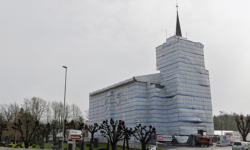 Kirke i gravlund innpakket i hvit- og blåstripet presenning, tårnspire stikker opp.